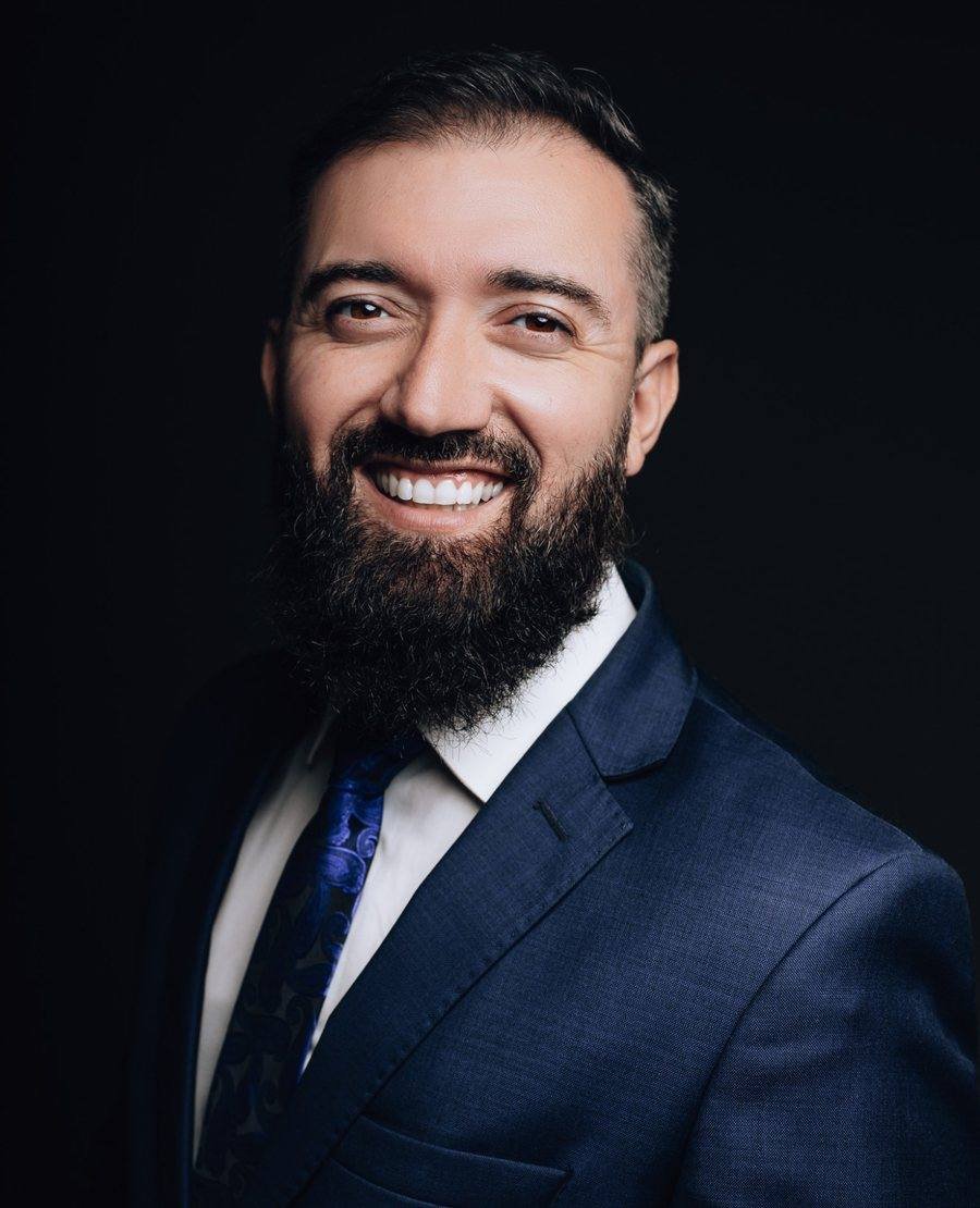 Denis Santos sorrindo, de terno azul-marinho e gravata